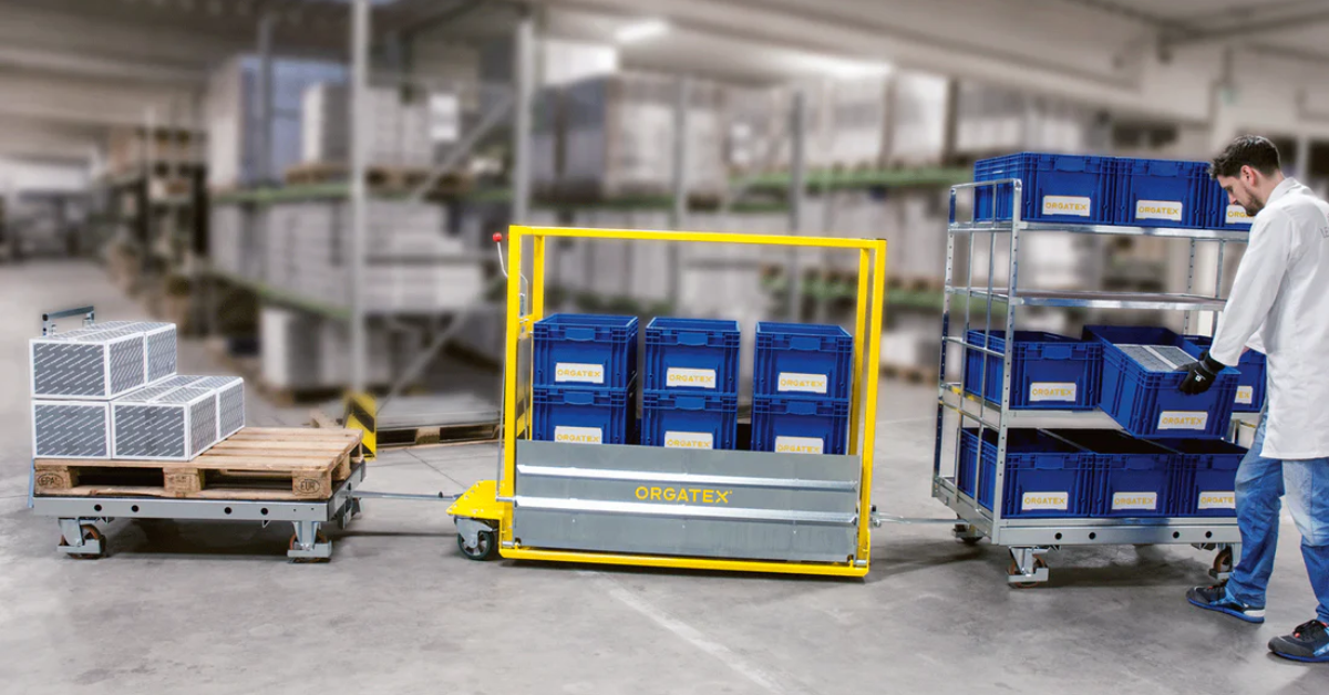 Carrinho logístico amarelo carregado de caixas azuis, com racks e um operador numa nave de armazenagem